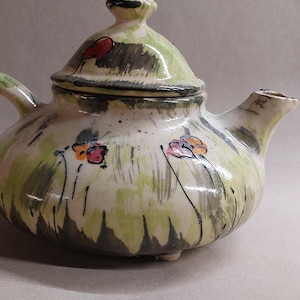 Könnte beinhalten: Eine handbemalte Keramik-Teekanne mit einer grün-weißen Glasur. Die Teekanne hat einen runden Körper und einen Deckel mit einem Knopf. Die Teekanne ist mit kleinen roten, orangefarbenen und gelben Blumen verziert.
