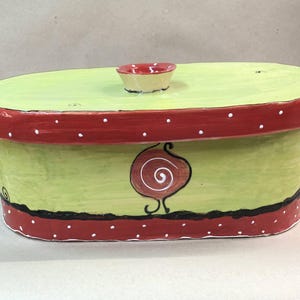 Può includere: Scatola decorativa ovale dipinta a mano con coperchio. La scatola è principalmente verde chiaro con bordo rosso e pois bianchi. Un disegno a spirale marrone è sul davanti. Il coperchio ha un piccolo pomello rotondo. Una linea nera ondulata con punti bianchi è in basso.