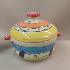 Peut inclure: Un plat à casserole en céramique colorée avec un couvercle. Le plat présente un design vibrant avec des bandes jaunes, oranges, bleues et blanches. Des poignées rouges en forme de champignon et un bouton de couvercle assorti ajoutent une touche fantaisiste.