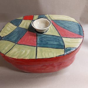 Könnte beinhalten: Eine handgefertigte Keramik-Servierschale mit Deckel. Die Schale ist rot mit einem Deckel mit einem farbenfrohen Patchwork-Design in Grün, Blau und Rot. Eine kleine, weiße Schale mit schwarzem Rand sitzt auf dem Deckel.