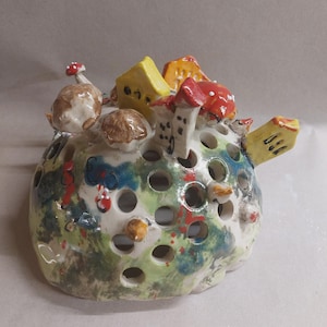 Könnte beinhalten: Eine skurrile Keramikskulptur mit einer perforierten, runden Form, bemalt mit einer Landschaft. Miniaturhäuser, Pilze und farbenfrohe Akzente in Rot, Gelb und Blau schmücken die Oberfläche. Ein einzigartiges Dekorationsstück.