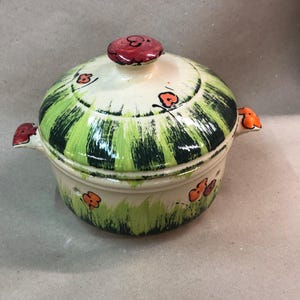 Peut inclure: Un plat à casserole en céramique avec couvercle, peint avec un motif fantaisiste. Le couvercle et le corps sont de couleur crème avec des rayures vertes verticales et des accents de fleurs orange. Les poignées et le bouton du couvercle sont également orange et rouge.