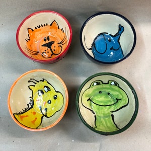 Könnte beinhalten: Vier Keramik-Schalen mit handbemalten Tiermotiven. Die Schalen sind beige mit orange, blau, gelb und grünen Akzenten. Die Tiere sind eine Katze, ein Elefant, eine Giraffe und ein Frosch.
