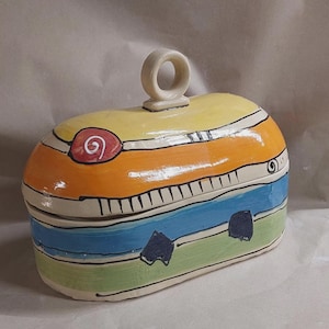 Könnte beinhalten: Eine handbemalte Keramikbox mit Deckel und Schlaufengriff. Die Box hat ein farbenfrohes Design mit Bändern in Gelb, Orange, Blau und Grün. Der Deckel hat ein rotes Spiralmuster. Die Box ist etwa 18 cm lang.