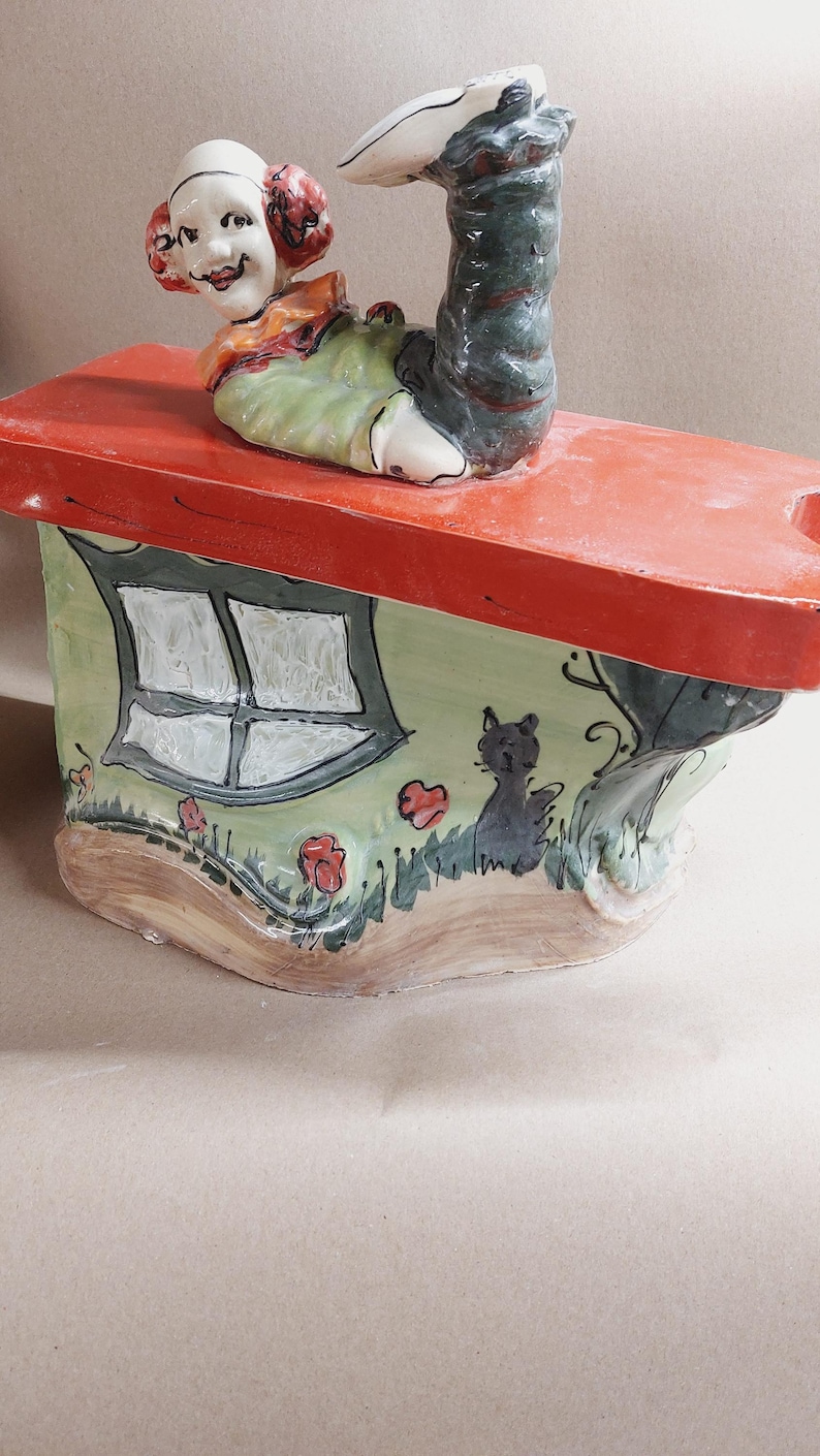 May include: A whimsical ceramic figurine featuring a smiling clown figure atop a miniature house. The house has a red roof, green walls, and a window. A black cat is painted on the side, with flowers and grass details.