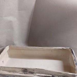 May include: A rectangular wooden box with a distressed, weathered finish. The box is painted in shades of white, grey, and blue, with visible wood grain. Decorative wooden cogs are affixed to the front. The interior is a light cream colour.
