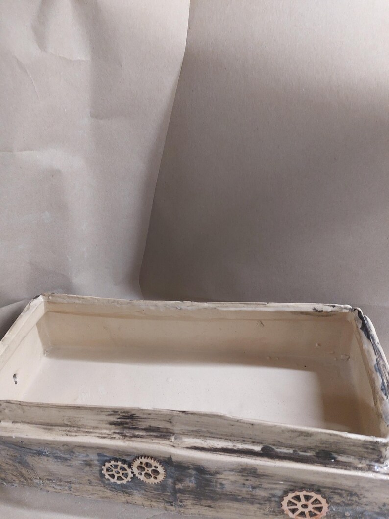 May include: A rectangular wooden box with a distressed, weathered finish. The box is painted in shades of white, grey, and blue, with visible wood grain. Decorative wooden cogs are affixed to the front. The interior is a light cream colour.
