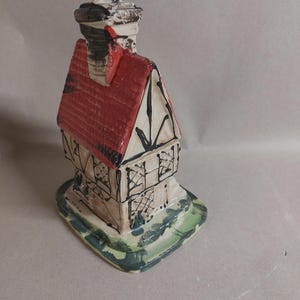 May include: A ceramic house figurine with a red roof, black chimney, and white and beige walls. The house has black trim and sits on a green and white base. The chimney is black and white.
