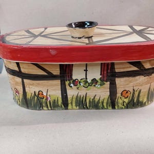 Peut inclure: Boîte à pain en céramique peinte à la main avec un couvercle. La boîte présente un bord rouge, un motif en bois brun clair et une petite poignée en forme de bol. Le devant de la boîte présente une scène peinte d'une maison avec des fleurs et de l'herbe.