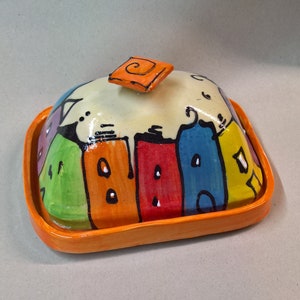 Könnte beinhalten: Eine farbenfrohe Butterdose aus Keramik mit einem skurrilen Design von Häusern, die in Grün-, Orange-, Rot-, Blau- und Gelbtönen bemalt sind. Die Schale hat einen orangefarbenen Rand und einen kleinen quadratischen Deckel mit einem Wirbelmuster.