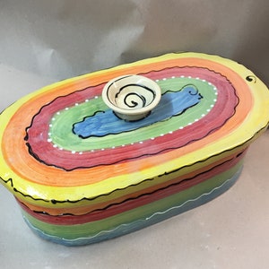 May include: A colourful, hand-painted ceramic dish with a lid. The oval dish has a rainbow design with layers of yellow, orange, red, green, and blue. A small, white bowl with a black spiral design sits on top of the lid.