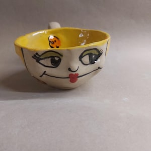 Könnte beinhalten: Handgefertigte Keramiktasse mit einem skurrilen Gesichtsdesign. Der Innenraum ist leuchtend gelb und die Außenseite zeigt ein gemaltes Gesicht mit schwarzen Augen, grünen Wimpern und roten Lippen. Ein kleiner Griff ist an der Seite angebracht.