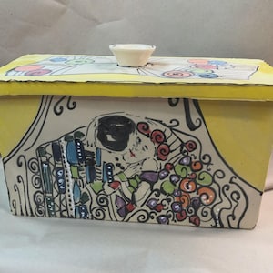 Può includere: Una scatola rettangolare in ceramica con coperchio e lati gialli. Il coperchio è decorato con vortici colorati e i lati presentano un dipinto di una coppia che si abbraccia. Un piccolo pomello bianco a forma di ciotola si trova sopra.