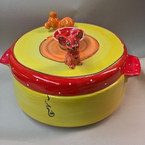 May include: A yellow ceramic casserole dish with a red rim and white polka dots. The lid features a small orange and red ceramic mouse figurine.