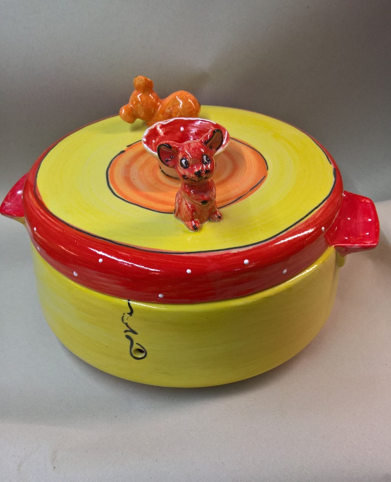 May include: A yellow ceramic casserole dish with a red rim and white polka dots. The lid features a small orange and red ceramic mouse figurine.