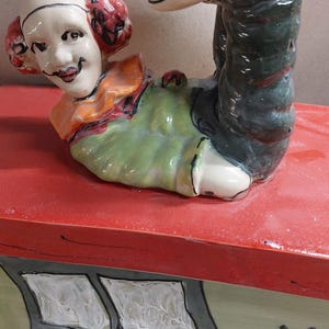 May include: A whimsical ceramic figurine of a clown in a playful pose, with red hair and a smiling face. The clown is atop a red surface, with a painted house design on the side, featuring a window and a cat.