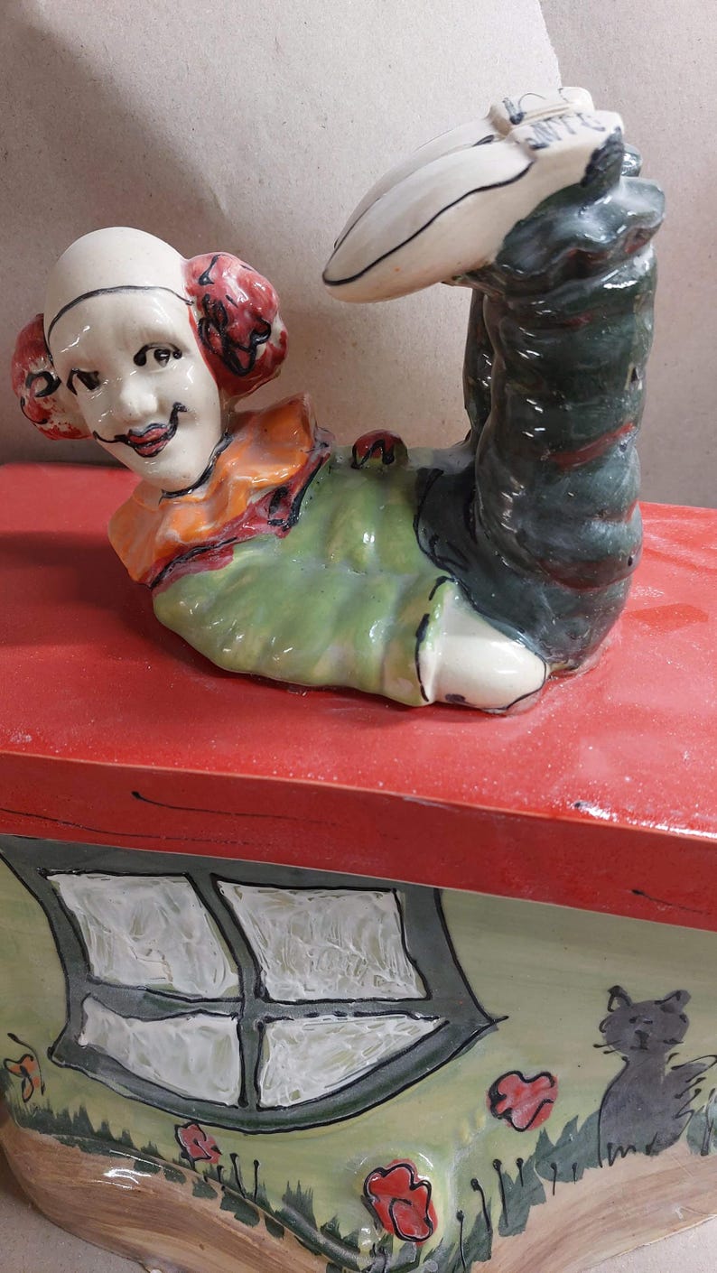 May include: A whimsical ceramic figurine of a clown in a playful pose, with red hair and a smiling face. The clown is atop a red surface, with a painted house design on the side, featuring a window and a cat.