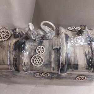 May include: A steampunk-inspired ceramic sculpture. It features a grey mouse figurine resting on a cylindrical form. The form is painted in grey and white, decorated with wooden gears and small mouse figurines.