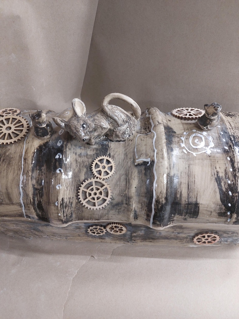 May include: A steampunk-inspired ceramic sculpture. It features a grey mouse figurine resting on a cylindrical form. The form is painted in grey and white, decorated with wooden gears and small mouse figurines.