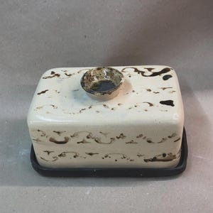 May include: A rectangular ceramic butter dish with a lid, featuring a cream-colored base adorned with dark brown abstract patterns. A small, round, decorative knob sits atop the lid. The dish rests on a black rectangular base.