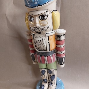 May include: A hand-painted ceramic nutcracker figurine with a blue hat, yellow beard, and gold glasses. The nutcracker is wearing a red and white striped jacket, green trousers, and blue boots with white polka dots. The figurine is standing on a blue base with white polka dots.