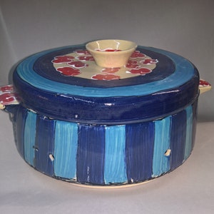 May include: A handmade ceramic casserole dish with a lid. The dish features a blue and white striped body, a blue lid with red floral accents, and a small beige bowl on top. The handles also have red floral accents.