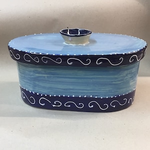 Puede incluir: Contenedor de cerámica ovalado pintado a mano con tapa. El contenedor está pintado en tonos azules con un borde azul oscuro y detalles de remolinos y puntos blancos. Un pequeño pomo redondo con borde azul se encuentra en la tapa.