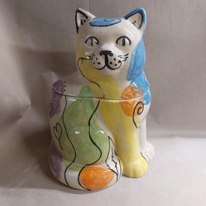 May include: A ceramic cat-shaped cookie jar. The jar is white with blue, yellow, green, orange, and purple painted accents. The cat has a smiling face and is sitting upright. The jar has a lid.