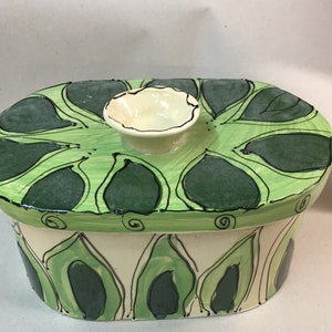 May include: Oval ceramic container with a lid, hand-painted with a green leaf design. The lid is light green with dark green leaf shapes outlined in black. The container's body is off-white with green leaf patterns. A small, off-white bowl sits atop the lid.