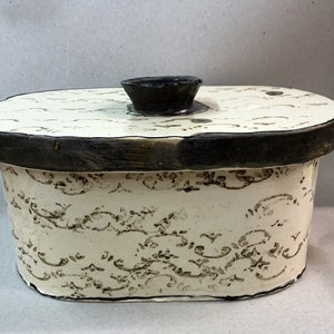 Peut inclure: Boîte ovale en céramique crème avec un bord et un couvercle marron foncé. Le couvercle a un petit bouton marron foncé. La boîte est décorée d'un motif orné répétitif marron foncé. Un récipient de rangement décoratif.