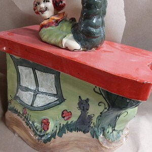 May include: A whimsical ceramic figurine of a clown figure upside down on a house-shaped base. The house has a red roof, green walls with a window, and a black cat illustration. The clown has a smiling face and is wearing a green outfit.