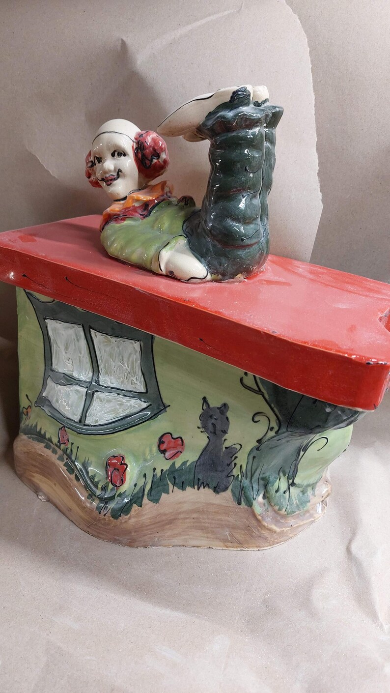 May include: A whimsical ceramic figurine of a clown figure upside down on a house-shaped base. The house has a red roof, green walls with a window, and a black cat illustration. The clown has a smiling face and is wearing a green outfit.