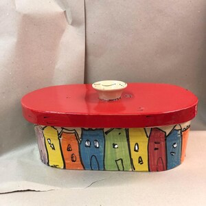May include: A hand-painted ceramic container with a red lid and a white knob. The container's base features a row of colorful houses in yellow, orange, blue, and green. The lid is oval-shaped.