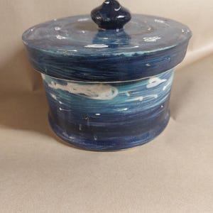 Puede incluir: Un tarro de cerámica hecho a mano con tapa, pintado en tonos azules y blancos. El tarro tiene un cuerpo cilíndrico y una tapa redondeada con un pequeño pomo azul oscuro. El diseño evoca un tema de cielo nocturno u océano.