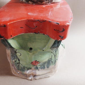 May include: A ceramic decorative piece featuring a red tabletop with a green base. The base is adorned with floral and leaf designs, and a small red flower. The legs of a figure are visible above the tabletop.