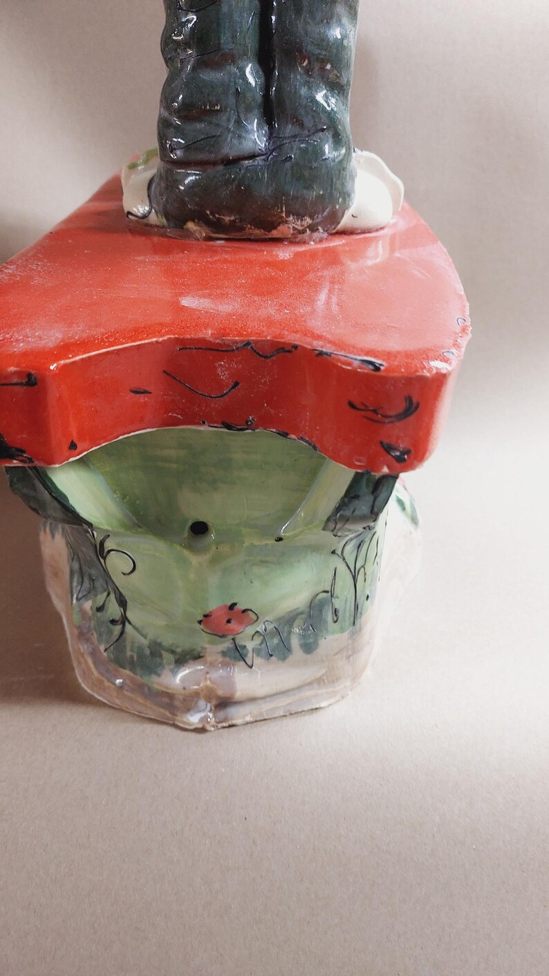 May include: A ceramic decorative piece featuring a red tabletop with a green base. The base is adorned with floral and leaf designs, and a small red flower. The legs of a figure are visible above the tabletop.