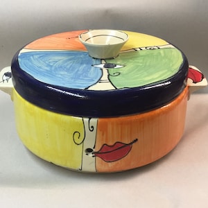 Può includere: Una casseruola in ceramica con coperchio. La casseruola è decorata con un design astratto colorato che presenta un viso con un occhio blu, labbra rosse e accenti gialli e arancioni. Il coperchio ha un piccolo pomo bianco.