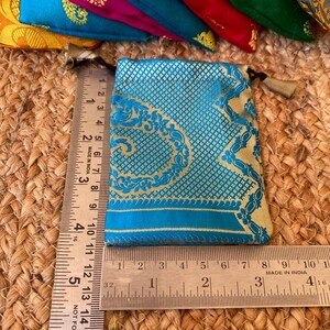 May include: A small, teal blue pouch with gold embroidery and a gold tassel. The pouch is made of a brocade fabric and is approximately 3.5 inches wide and 4 inches tall.