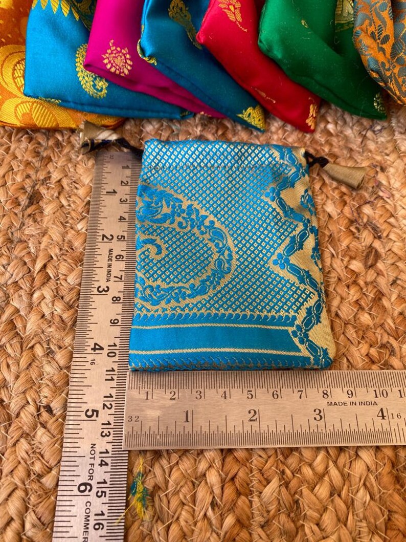 May include: A small, teal blue pouch with gold embroidery and a gold tassel. The pouch is made of a brocade fabric and is approximately 3.5 inches wide and 4 inches tall.