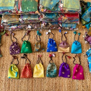 May include: Assortment of small drawstring bags made from colorful patterned fabric. The bags are arranged in two rows on a woven surface. The bags are all different colors and patterns, including purple, green, blue, yellow, and red.