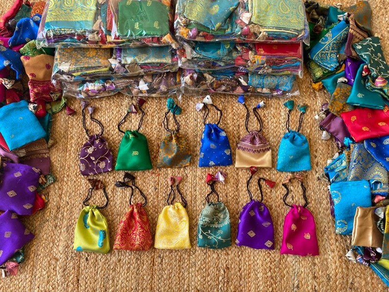 May include: Assortment of small drawstring bags made from colorful patterned fabric. The bags are arranged in two rows on a woven surface. The bags are all different colors and patterns, including purple, green, blue, yellow, and red.
