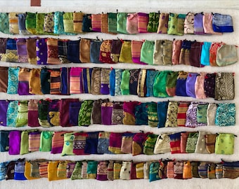 May include: A collection of small, colorful fabric pouches arranged on a white woven surface. The pouches are made from various patterned fabrics in shades of green, purple, gold, and blue. Each pouch has a drawstring closure. The overall aesthetic is bohemian and handcrafted.