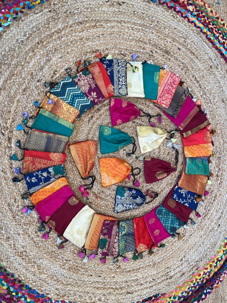May include: A collection of 20 colorful fabric pouches with tassels. The pouches are arranged in a circle on a woven rug. The pouches are made of different fabrics, including silk, cotton, and velvet. They are decorated with various patterns, including floral, geometric, and paisley.