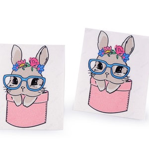 Op de afbeelding: Twee witte stickers met een cartoon afbeelding van een konijn dat een blauwe bril en een bloemenkrans draagt. Het konijn zit in een roze zak.