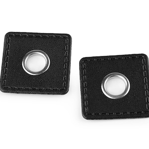 Könnte beinhalten: Zwei schwarze Lederquadrate mit silbernen Ösen in der Mitte. Die Quadrate haben einen genähten Rand.