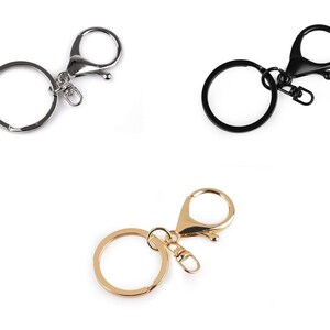 Könnte beinhalten: Drei Schlüsselanhänger in Silber, Schwarz und Gold vor weißem Hintergrund. Jeder Anhänger hat einen runden Ring und einen Karabinerhaken. Geeignet zum Befestigen von Schlüsseln oder anderen kleinen Gegenständen.