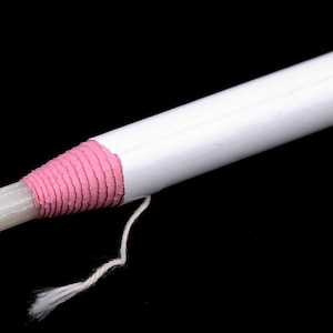 May include: Close-up of a white candle with a pink, wrapped section near the top. A white wick extends from the candle, with a small, frayed end. The background is solid black.