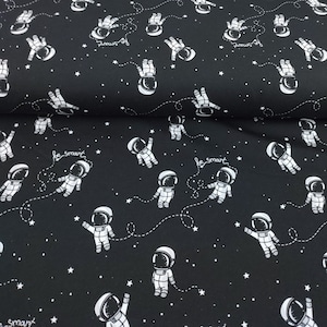 Op de afbeelding: Zwarte stof met een wit, herhalend patroon van astronauten in de ruimte. De astronauten dragen ruimtepakken en hebben een stervormig ontwerp op hun helmen. De stof heeft een witte tekst die "be smart" en "dreams of" zegt.