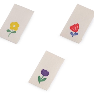 Op de afbeelding: Drie rechthoekige crèmekleurige stoffen labels, elk met een ander bloemenontwerp. Eén heeft een gele bloem, een andere een rode tulp en de derde een paarse bloem. Allemaal hebben ze groene stelen en bladeren.