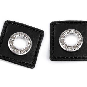 Op de afbeelding: Twee zwarte vierkante gordijnbandjes met zilveren oogjes versierd met heldere strass steentjes.